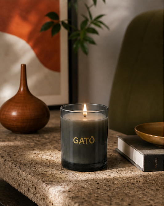 A dark gray glass jar candle with a gold 'GATO' logo sits lit on a stone surface in a styled interior setting with a wooden vase, framed art, and a green chair in the background.
