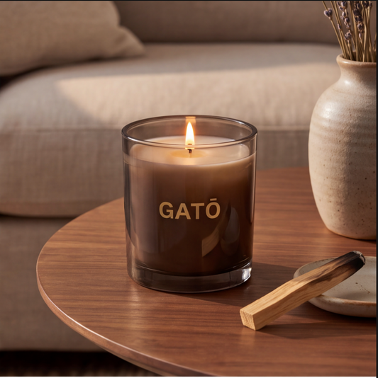 A lit candle in a dark smoky glass jar with the word 'GATO' printed in gold on the front, set on a round wooden table in a cozy living room scene with a vase and matchstick.