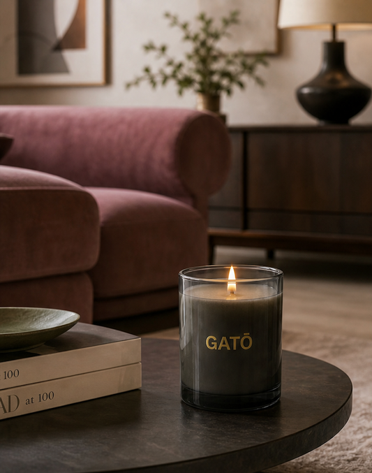 A lit GATO candle in a clear glass jar sits on a dark round table in a cozy living room setting with a pink sofa, books, and a lamp in the background.