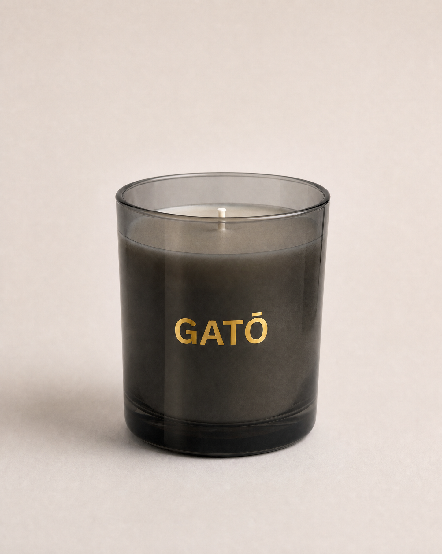 A dark gray glass jar candle with the brand name 'GATŌ' printed in gold lettering on the front, set against a neutral light beige background.