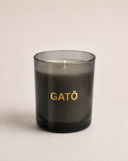 A dark gray glass jar candle with the brand name 'GATŌ' printed in gold lettering on the front, set against a neutral light beige background.