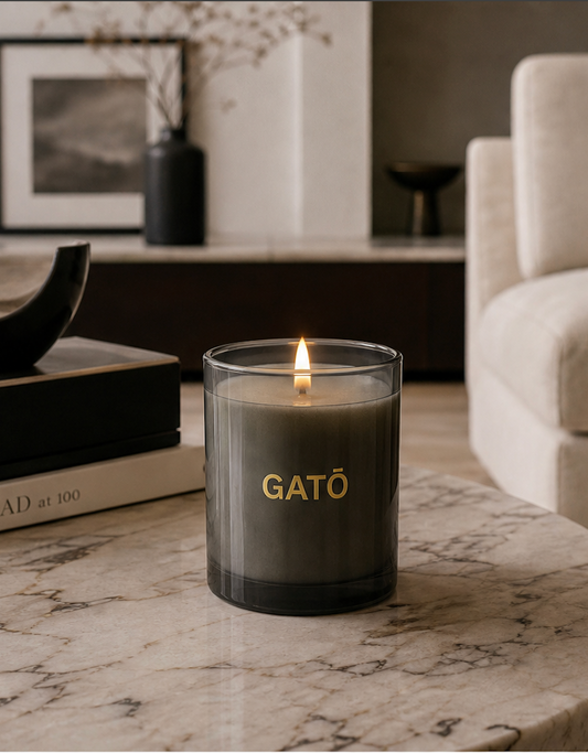 A lit GATŌ candle in a dark gray glass jar sits on a marble table in a modern living room setting.