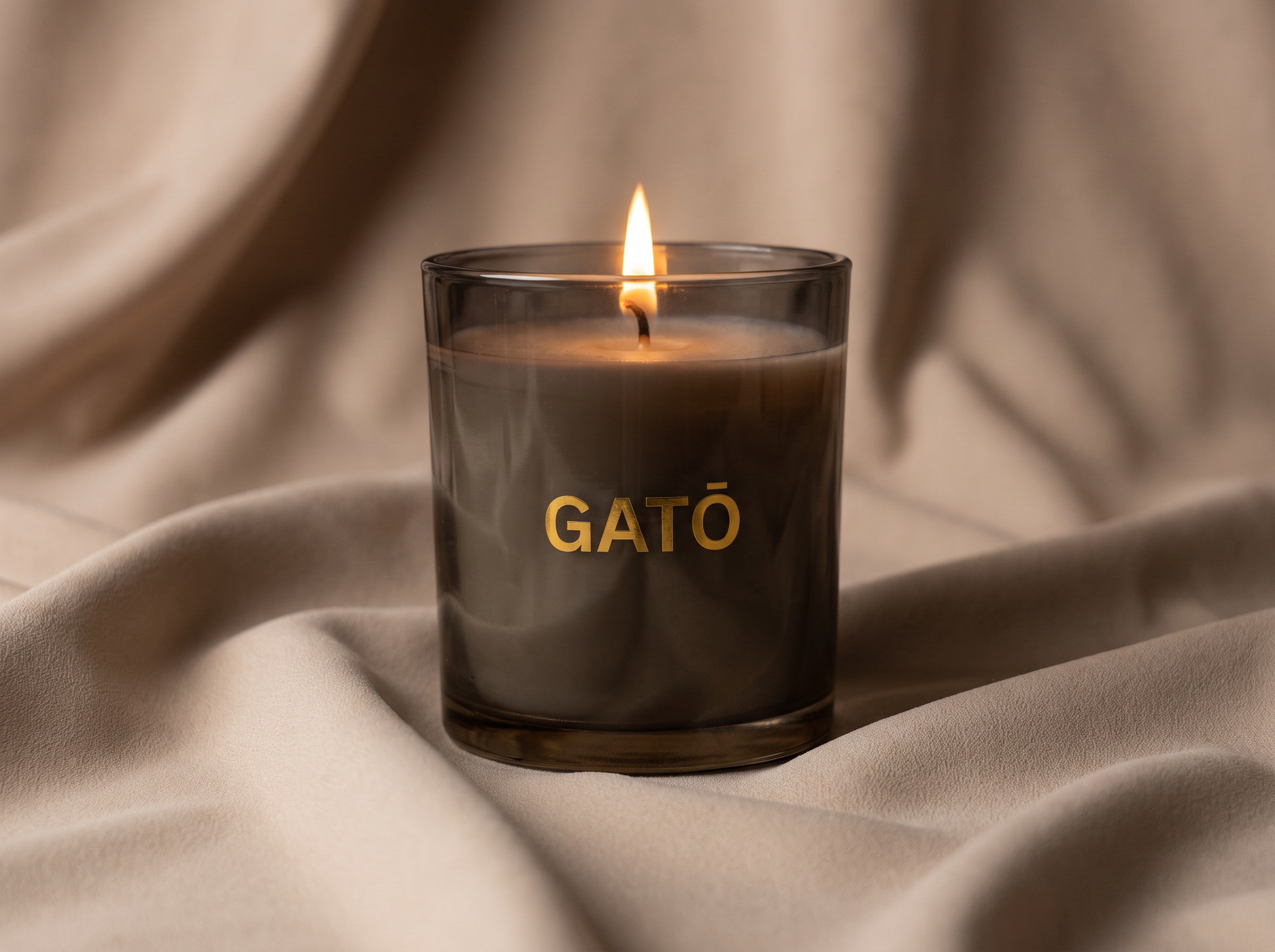 A lit candle in a dark glass jar with the brand name 'GATO' printed in gold on the front, set against a draped beige fabric background.
