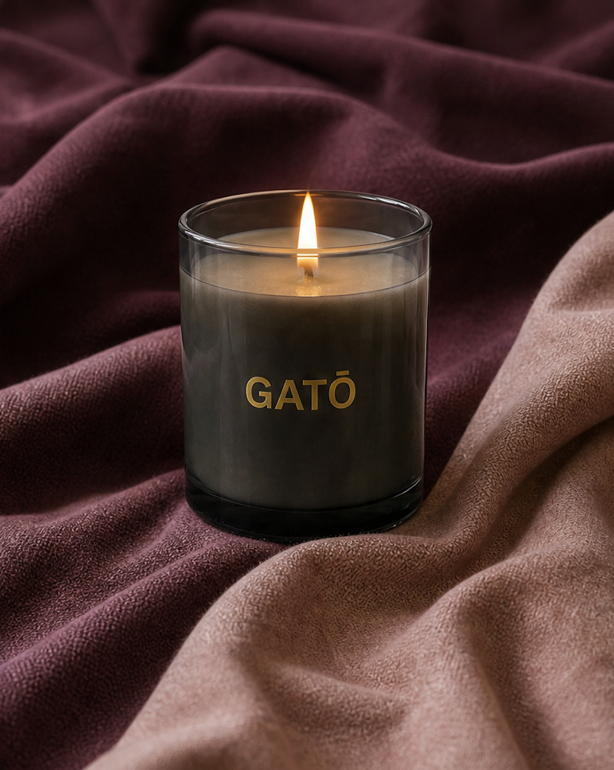 A lit candle in a clear glass jar with the brand name GATÓ printed in gold on the front, set against a backdrop of draped mauve and dusty rose fabric.