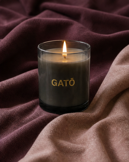 A lit candle in a clear glass jar with the brand name GATÓ printed in gold on the front, set against a backdrop of draped mauve and dusty rose fabric.