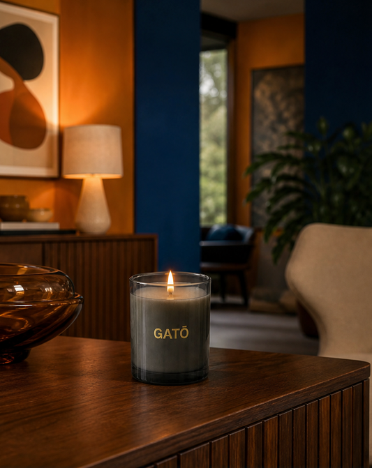 A lit GATO candle in a clear glass jar sits on a dark wood table in a warmly lit modern living room with orange and blue walls.