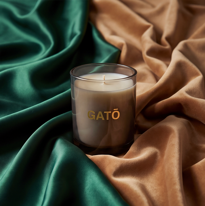 A clear glass jar candle with cream wax and a gold 'GATŌ' logo, styled on draped emerald green and tan silk fabrics.